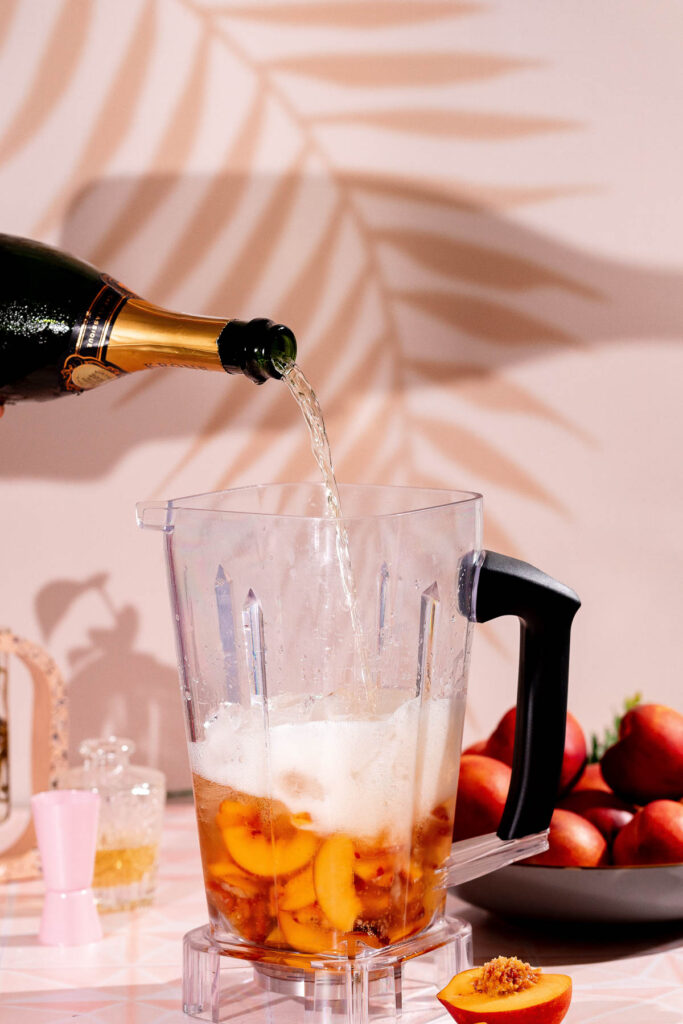 Prosecco is being poured into a blender with sliced peaches, while a bowl of peaches and kitchen items are visible in the background.