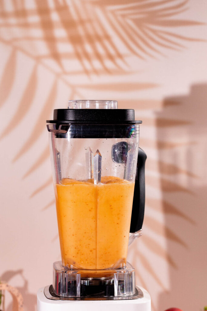 A blender filled with peach bellini sits on a countertop, casting palm leaf shadows on the light-colored wall.