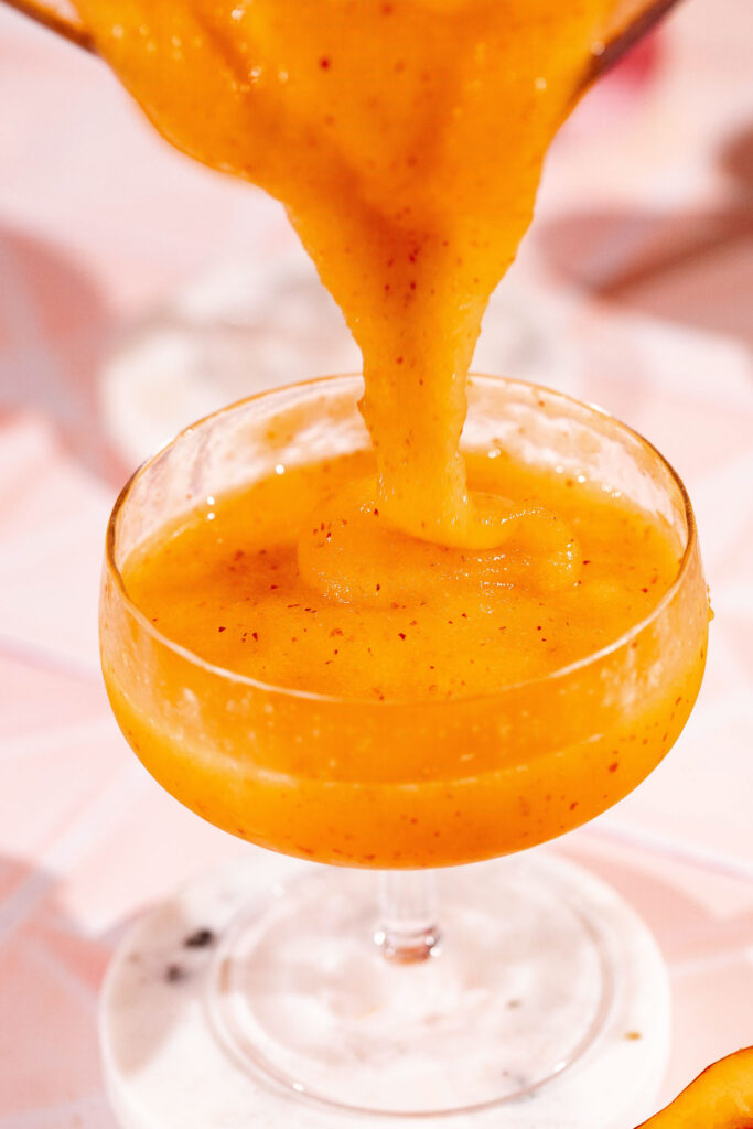 A thick peach bellini with specks is poured into a clear glass on a pink surface.