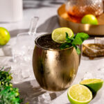 A Kentucky Mule garnished with lime and mint is accompanied by lime slices, ice cubes, and a glass decanter in the background.