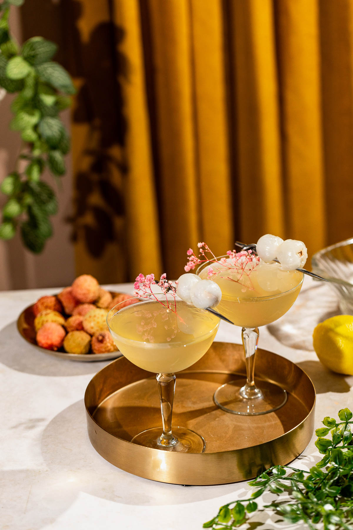 Two lychee martinis garnished with lychees and pink flowers sit on a brass tray, with a plate of lychees and a lemon in the background.