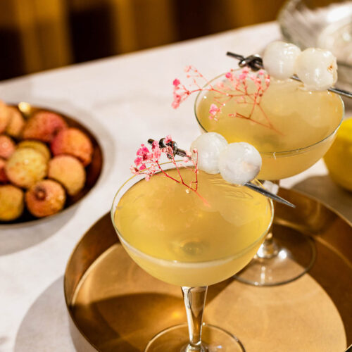 Two lychee martini cocktails with white berries and pink flowers on a gold tray are set against a background featuring a bowl of lychees on a white surface.