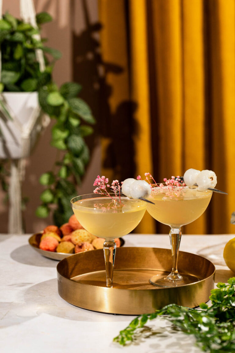 Two lychee martini cocktails garnished with lychee fruit and pink flowers sit on a brass tray against a backdrop of yellow curtains, plants, and a bowl of lychees.