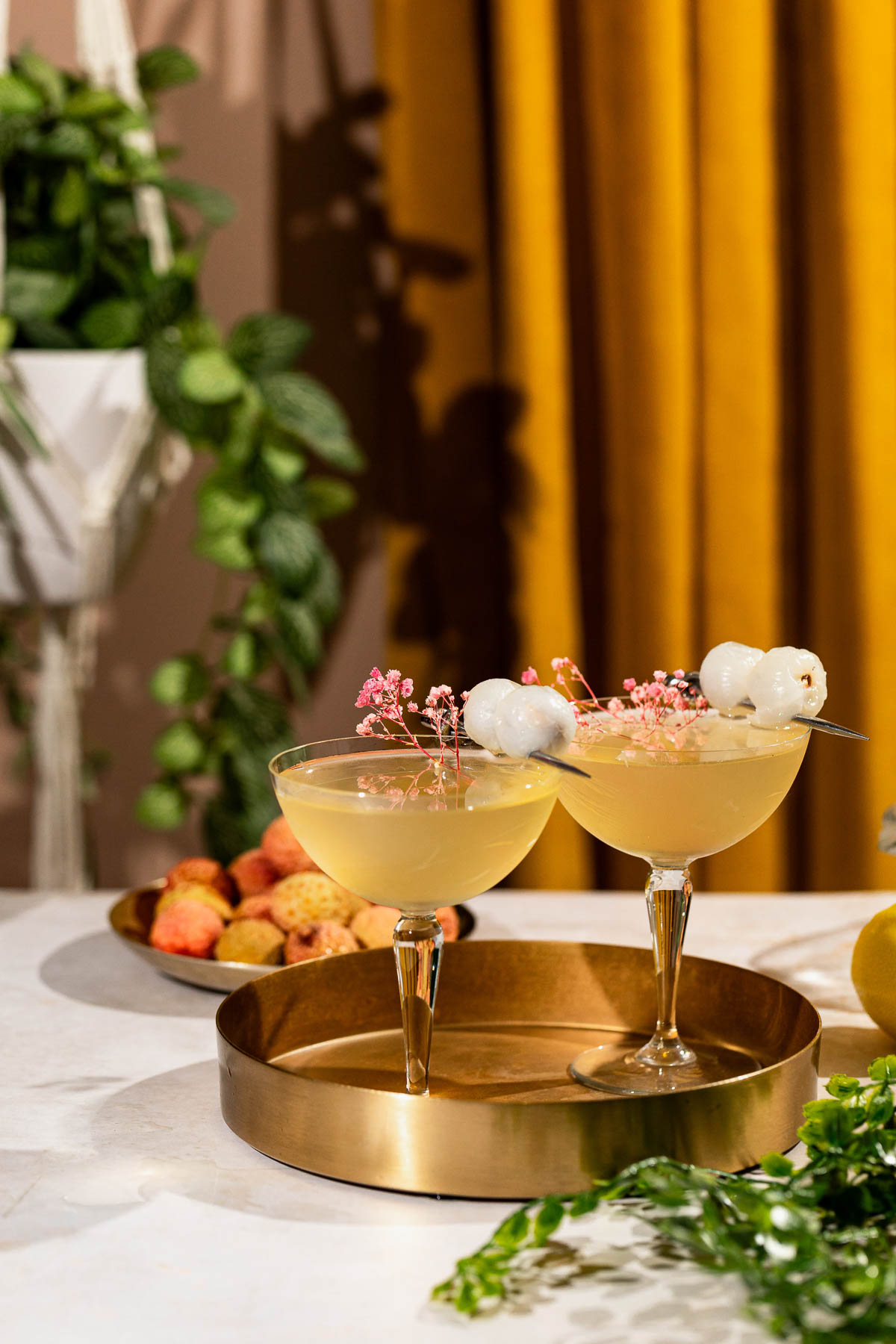 Two lychee martini cocktails garnished with lychee fruit and pink flowers sit on a brass tray against a backdrop of yellow curtains, plants, and a bowl of lychees.