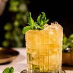 A glass of mint julep iced cocktail, garnished with mint leaves, sits on a coaster surrounded by ice and additional mint.