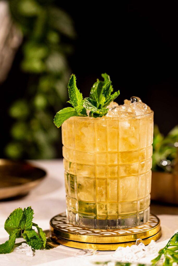 A glass of mint julep iced cocktail, garnished with mint leaves, sits on a coaster surrounded by ice and additional mint.