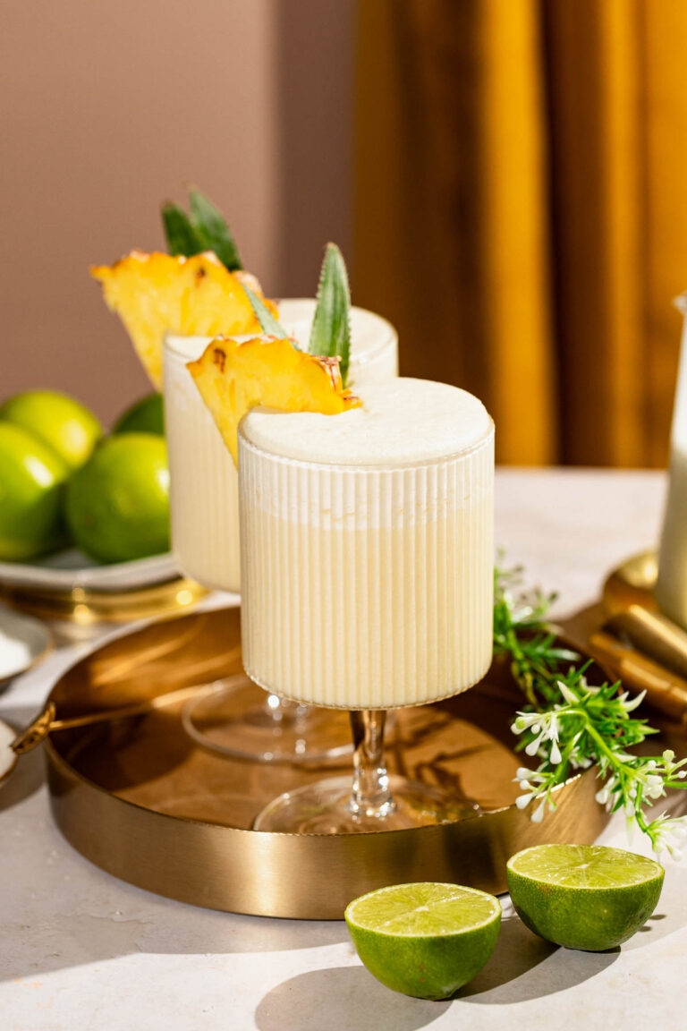 Two frothy pina colada cocktails garnished with pineapple slices sit on a gold tray surrounded by fresh limes and flowers.