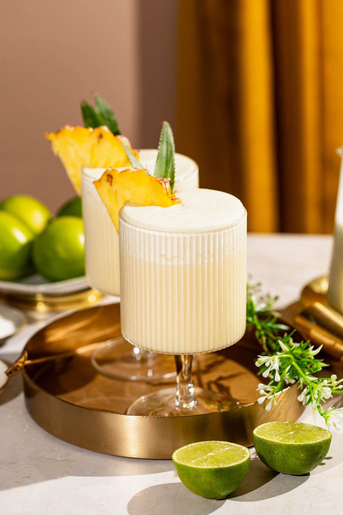 Two frothy pina colada cocktails garnished with pineapple slices sit on a gold tray surrounded by fresh limes and flowers.
