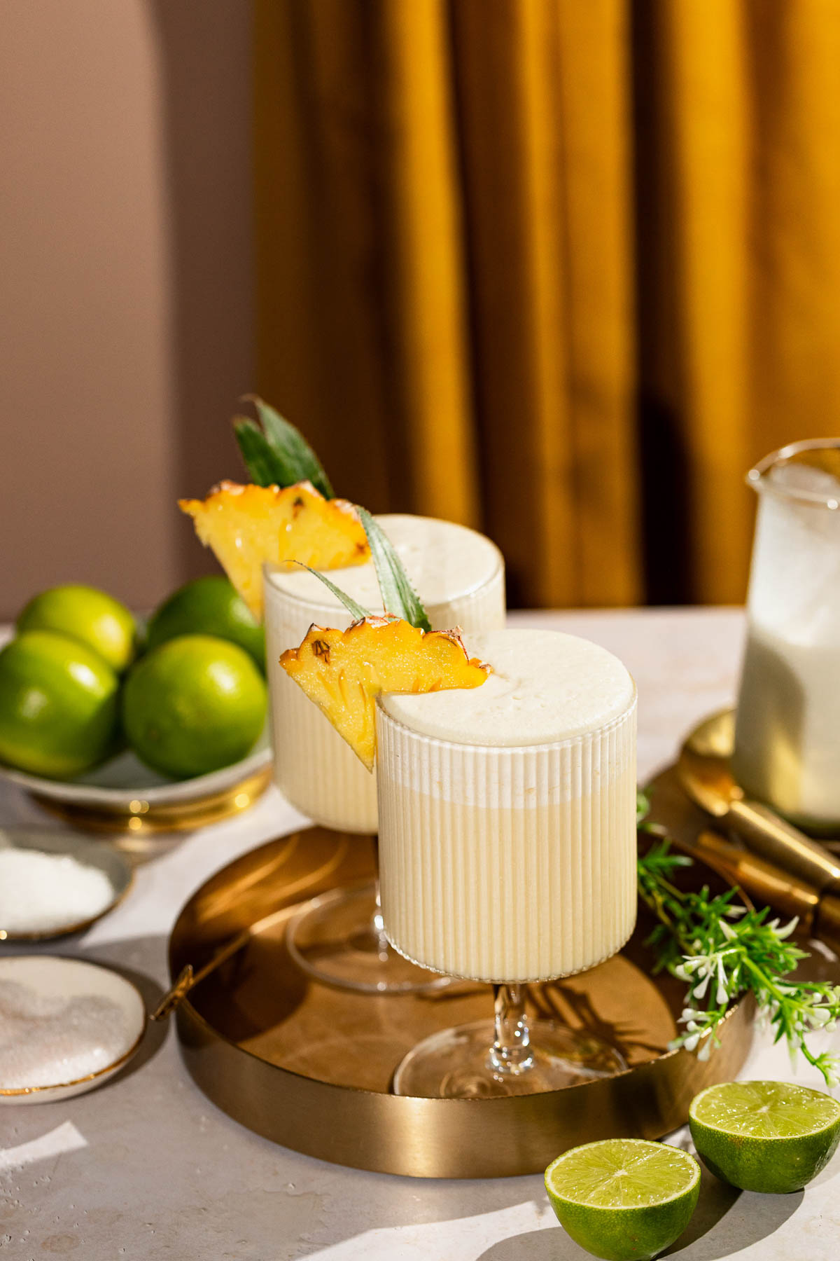 Two creamy pina coladas garnished with pineapple slices sit on a gold tray surrounded by limes, sugar, and a pitcher, with a yellow curtain in the background.