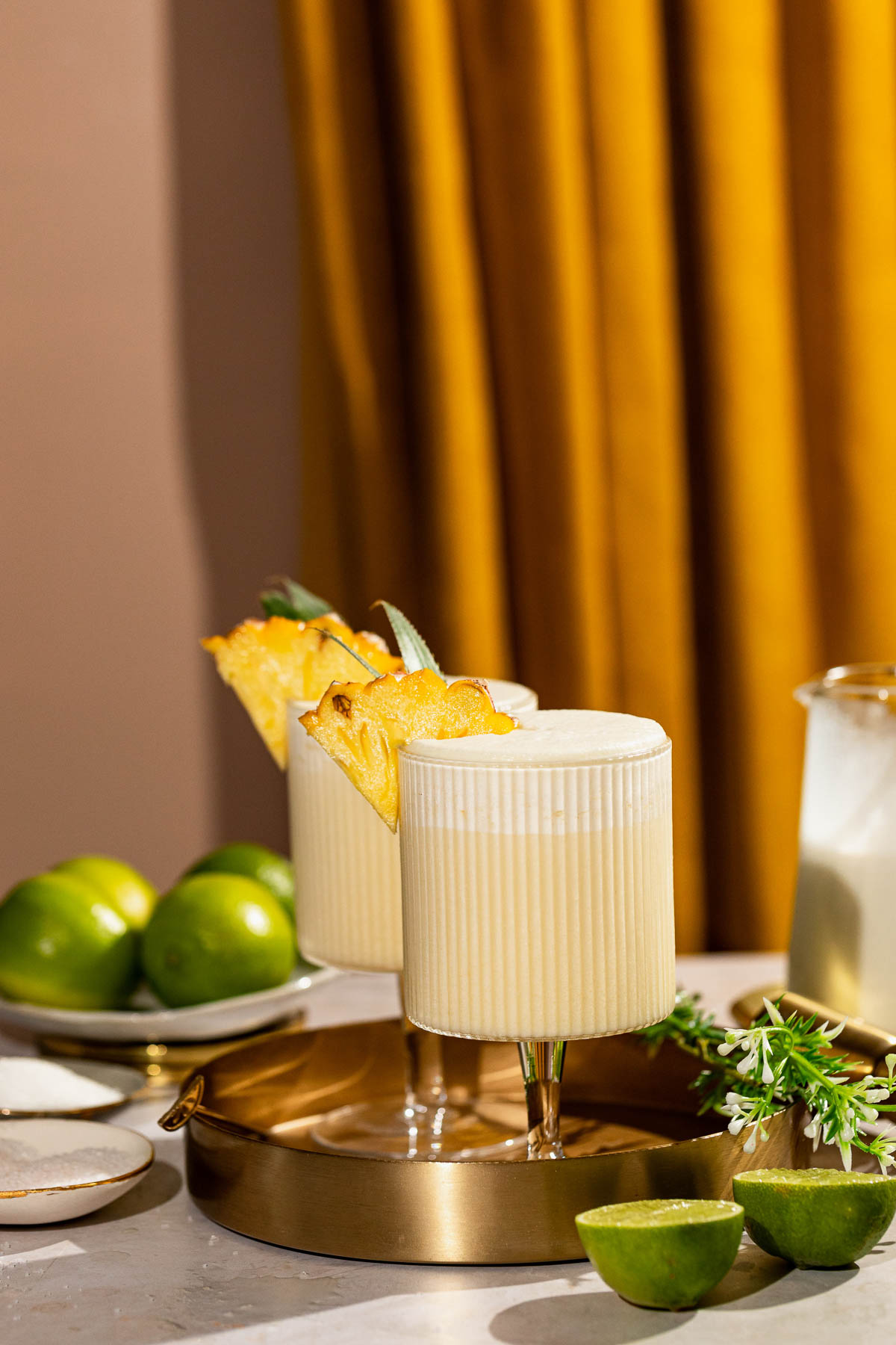 Two creamy pina colada cocktails with pineapple garnishes are on a gold tray with limes nearby, set against a mustard-colored curtain.