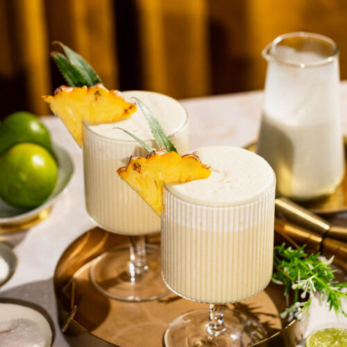 Two piña coladas garnished with pineapple slices sit on a gold tray, accompanied by lime halves and a pitcher, with a yellow curtain in the background.