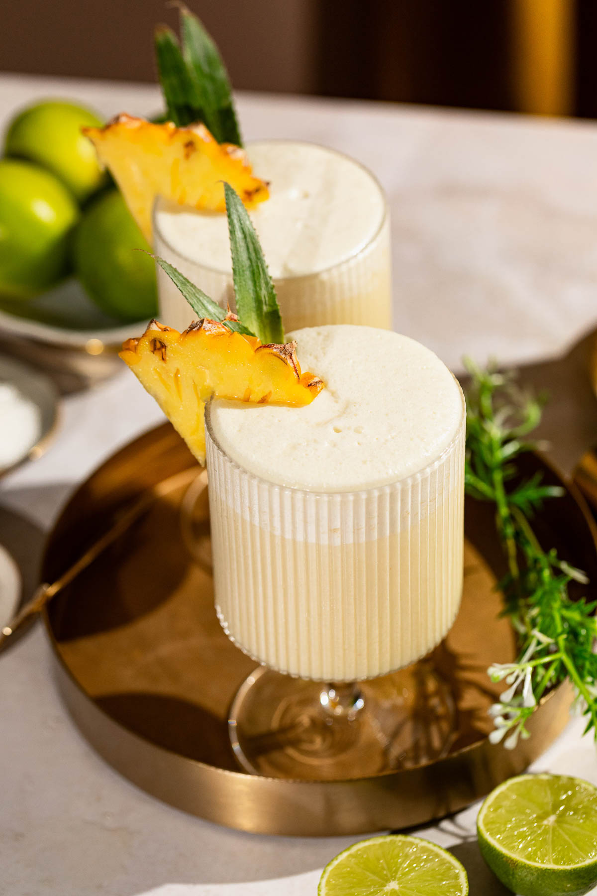 Two frothy pina coladas in ribbed glasses garnished with pineapple slices are on a decorative tray alongside lime halves and whole limes.