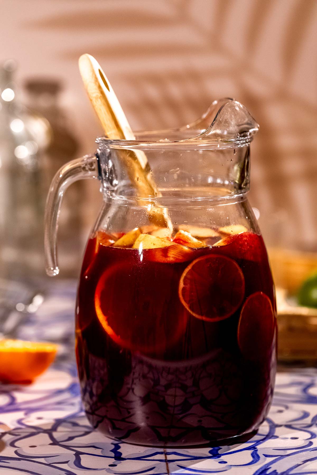 A glass pitcher of sangria with sliced citrus and ice, containing a wooden spoon, rests on a patterned surface.