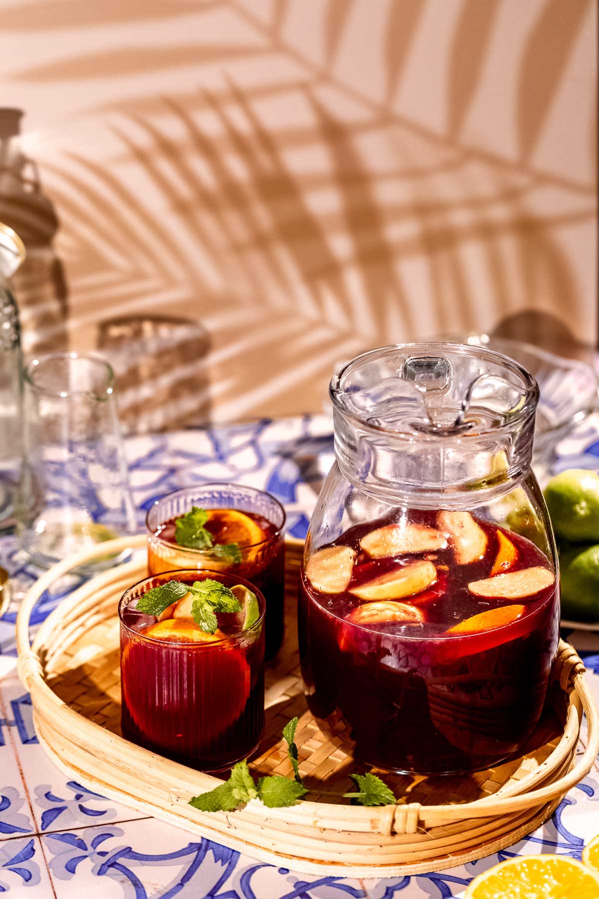 A glass pitcher and two glasses of red sangria with fruit and mint rest on a woven tray atop tiles, with limes and patterned shadows behind.