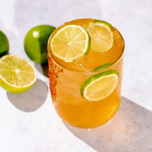 A glass of iced Cadillac margarita drink with a chili powder rim and lime slices is surrounded by whole and sliced limes on a light surface.