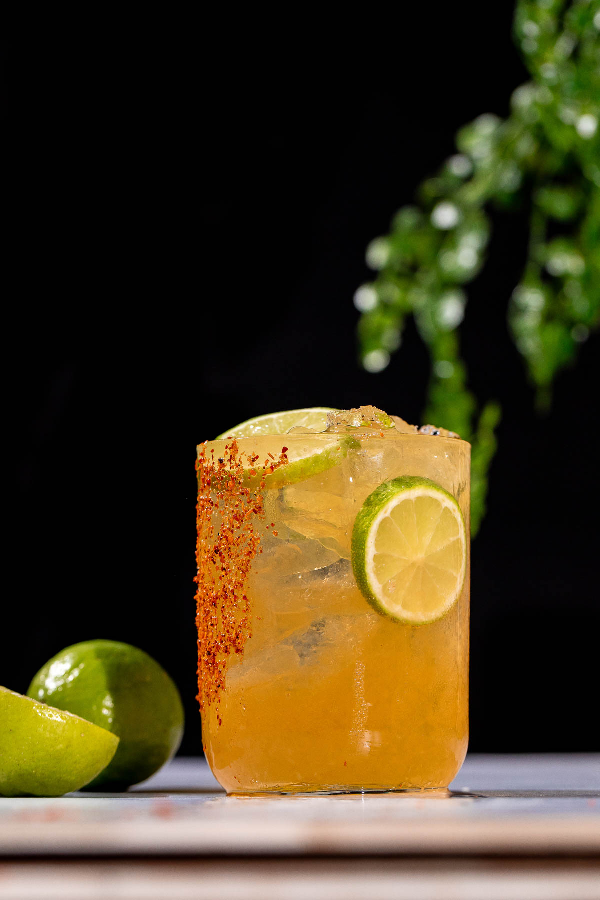 An orange Cadillac margarita cocktail with ice, lime slices, and chili powder on the rim sits on a table beside fresh limes, with a blurred plant in the background.