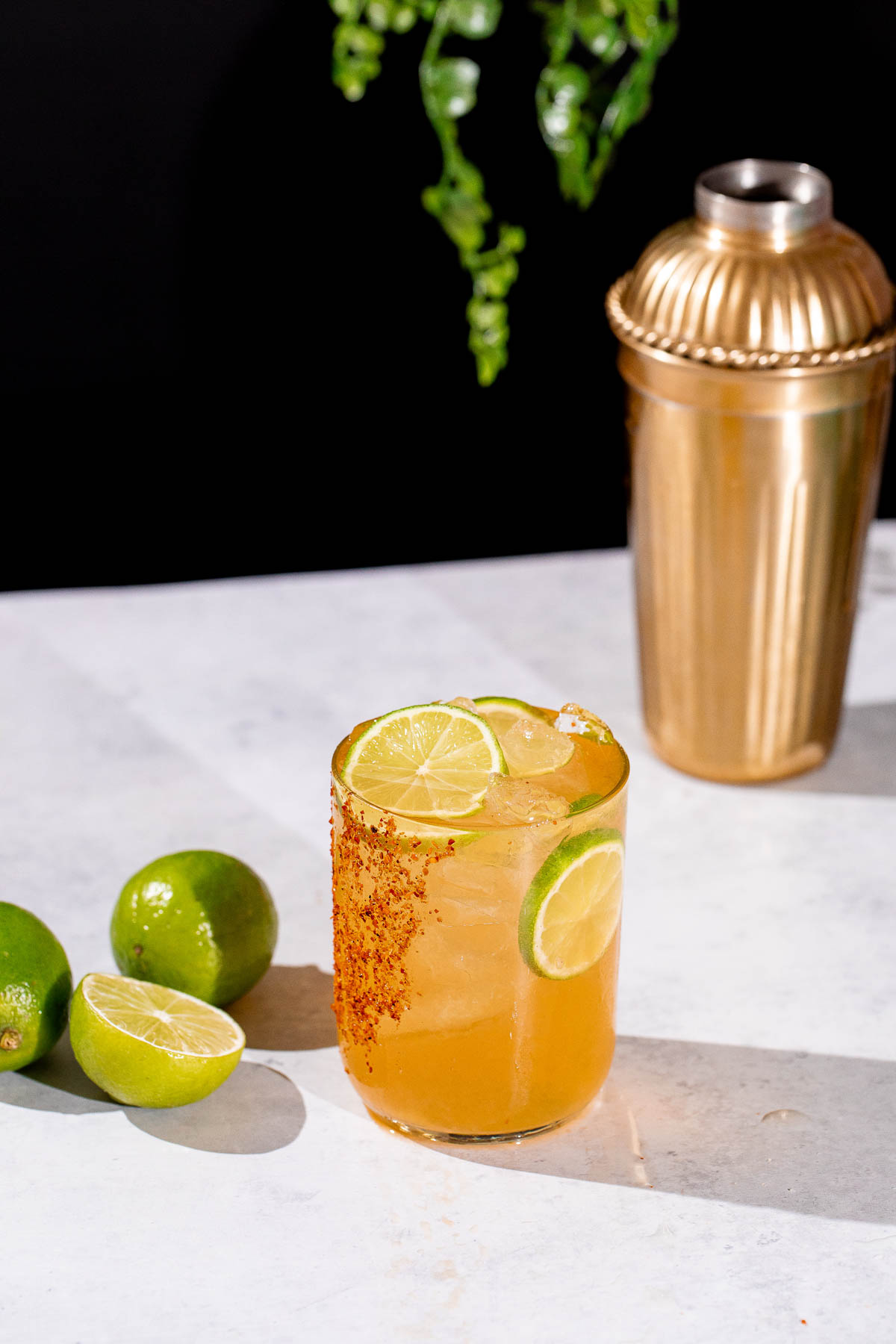 A chili-rimmed cadillac margarita of cocktail with ice and lime slices sits beside whole and halved limes and a gold shaker on a white surface.