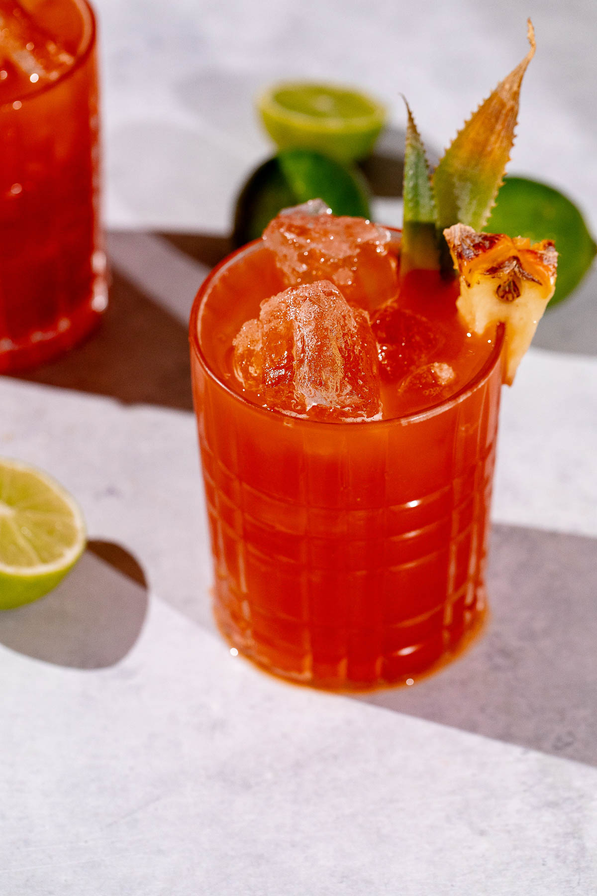A Jungle Bird cocktail with ice, garnished with pineapple and leaves, sits on a light surface beside halved limes.