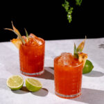 Two Jungle Bird cocktails with pineapple garnishes and ice sit on a light surface beside halved limes, set against a black background with greenery overhead.