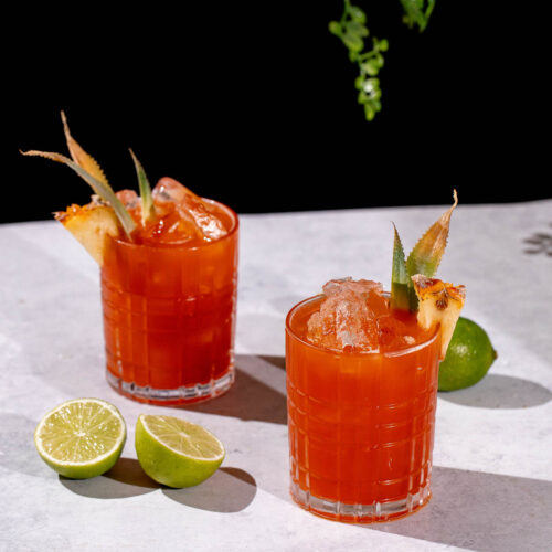 Two Jungle Bird cocktails with pineapple garnishes and ice sit on a light surface beside halved limes, set against a black background with greenery overhead.