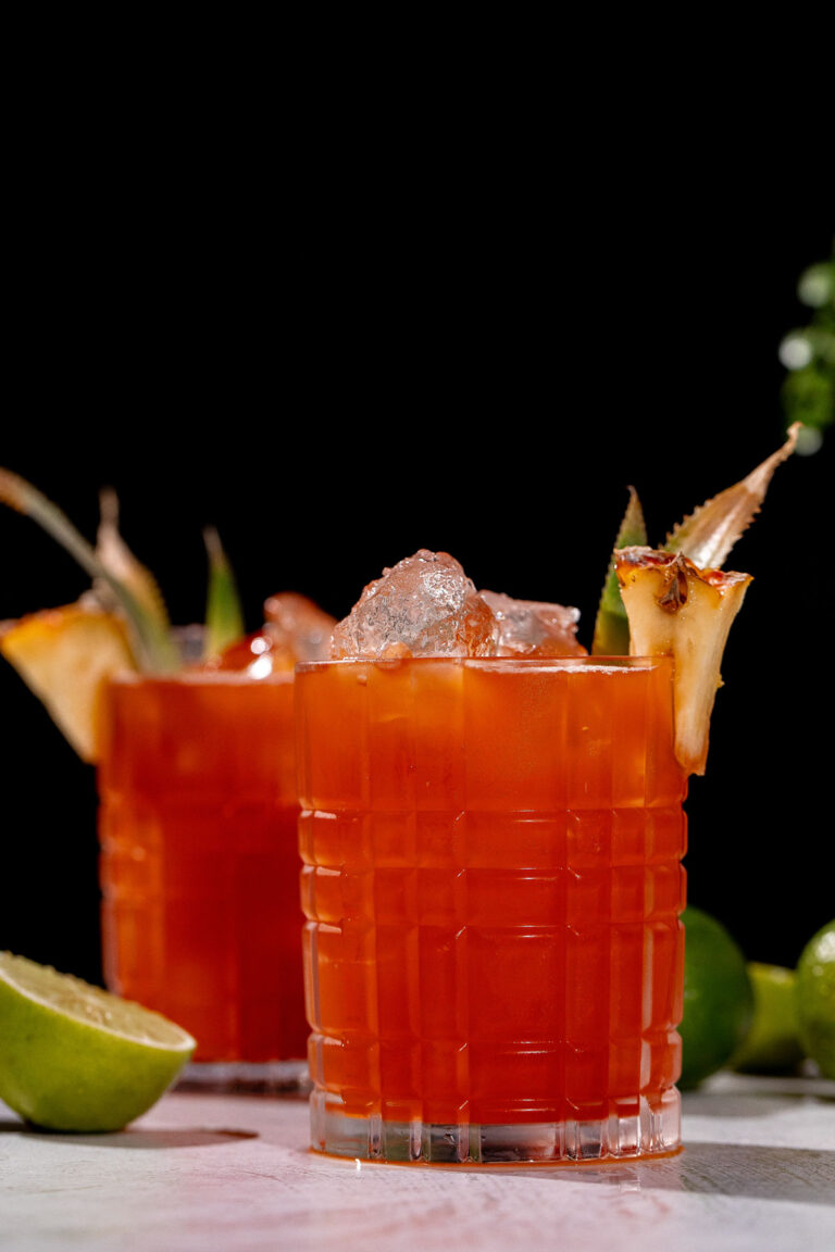 Two Jungle Bird cocktails with ice and pineapple wedges sit on a light surface, with limes behind them against a black background.