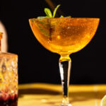 A coupe glass of sparkling orange old cuban cocktail with mint garnish sits on a table, blurred drink and lime in the background.