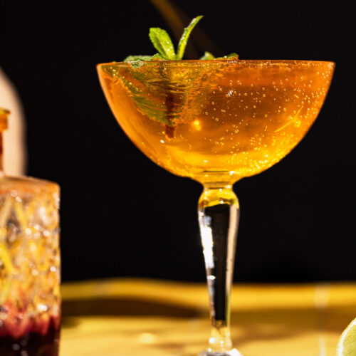A coupe glass of sparkling orange old cuban cocktail with mint garnish sits on a table, blurred drink and lime in the background.
