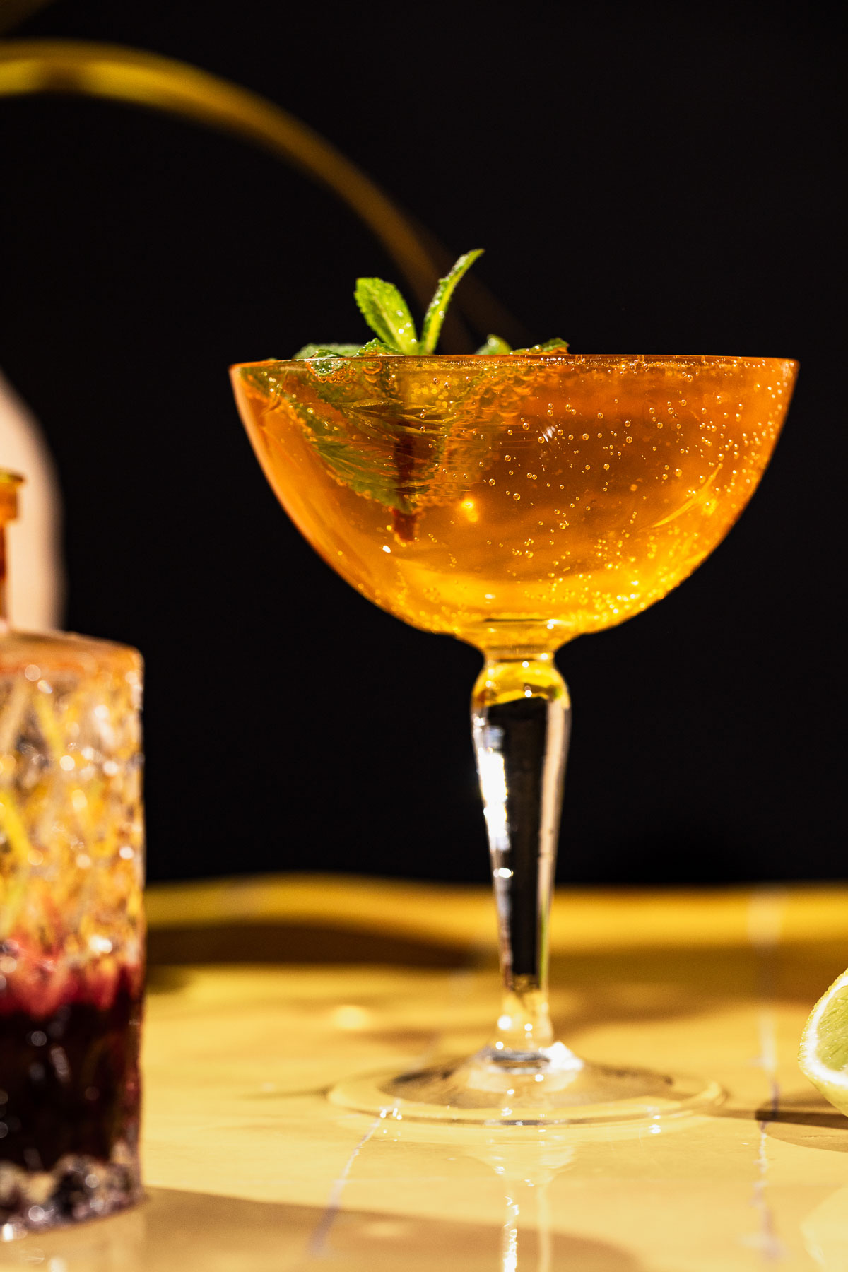 A coupe glass of sparkling orange old cuban cocktail with mint garnish sits on a table, blurred drink and lime in the background.