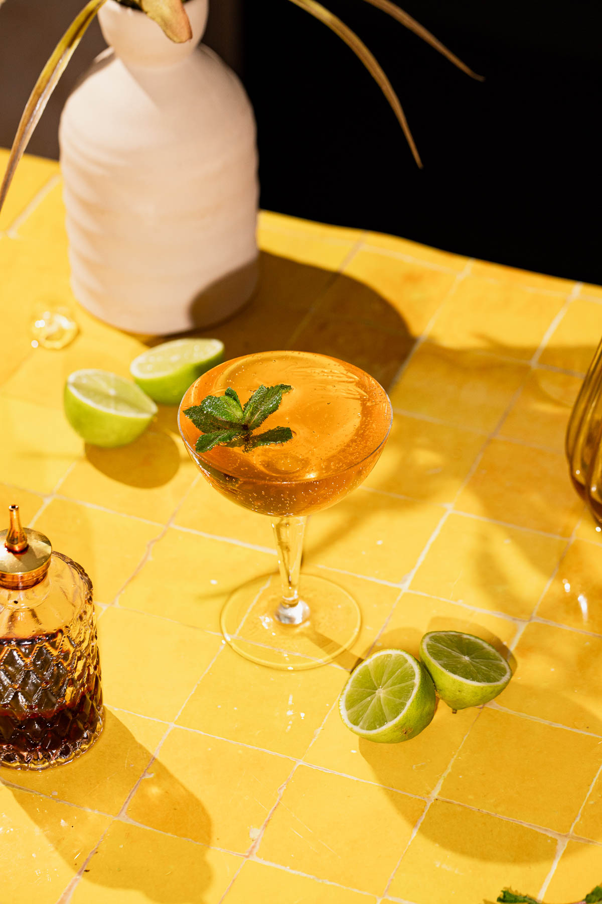 A mint-garnished old cuban cocktail sits on a yellow tiled table with halved limes, a glass bottle, and a white vase of green leaves.