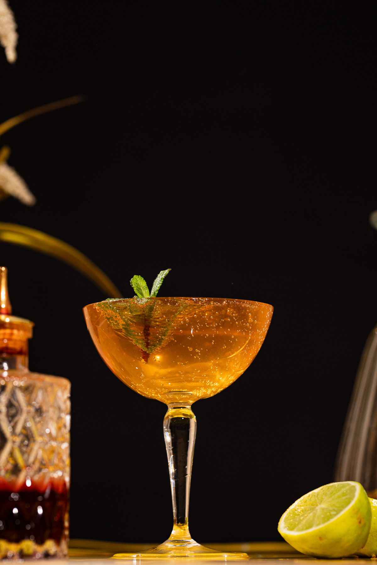 Coupe glass with an old cuban cocktail and mint, beside halved lime and decorative bottle on a black background.