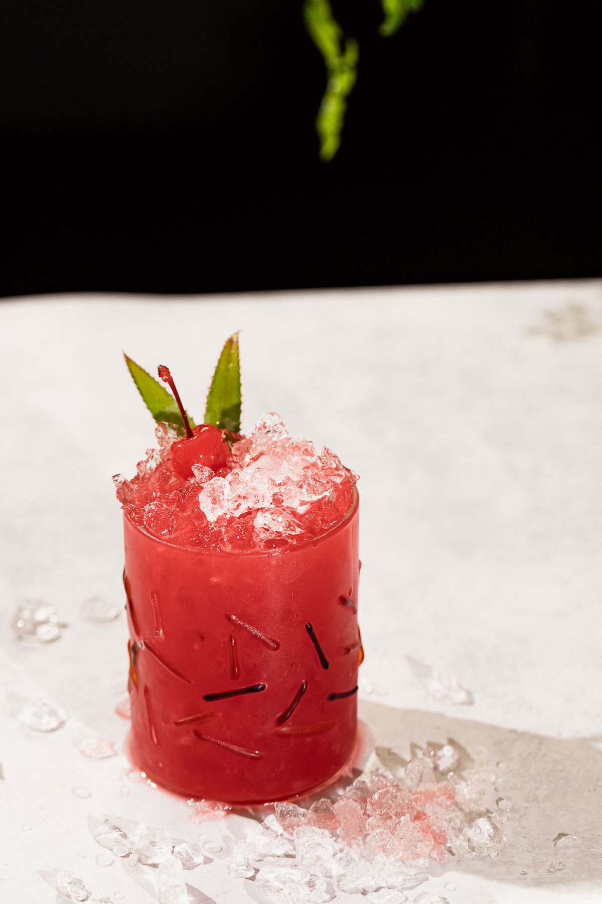 A red rum runner cocktail with crushed ice, topped with a cherry and green leaves, sits on a white surface scattered with ice pieces.