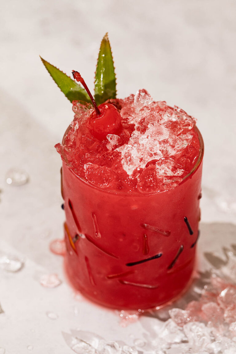 A rum runner cocktail with crushed ice, a maraschino cherry, and green leaves in a clear glass on a light surface.