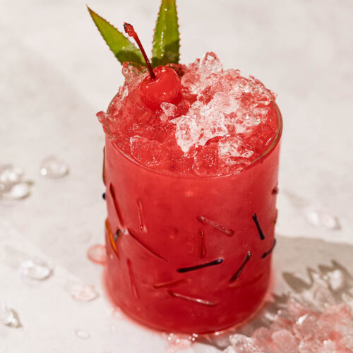 A red rum runner cocktail garnished with a cherry and two green leaves sits on a light surface amid scattered ice.