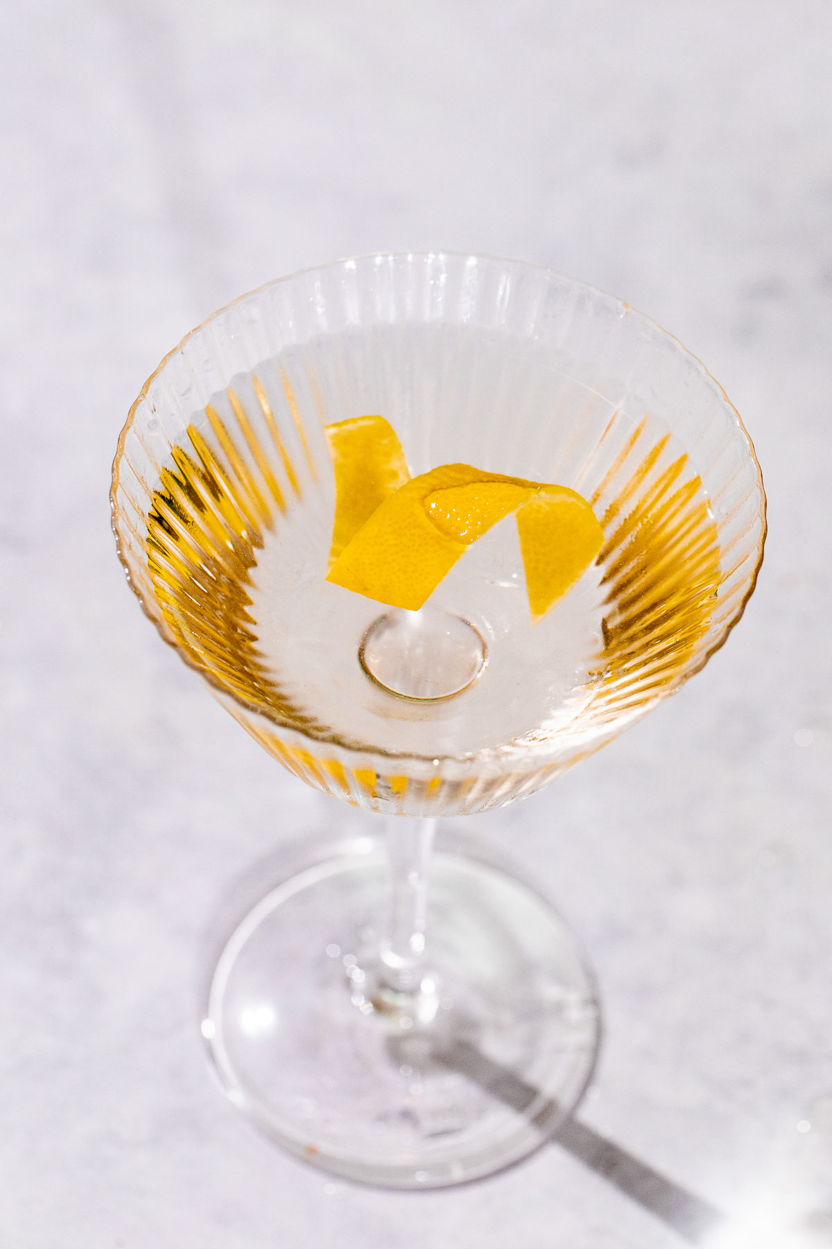 A clear vesper martini with a lemon twist is served in a stemmed glass on a light surface.