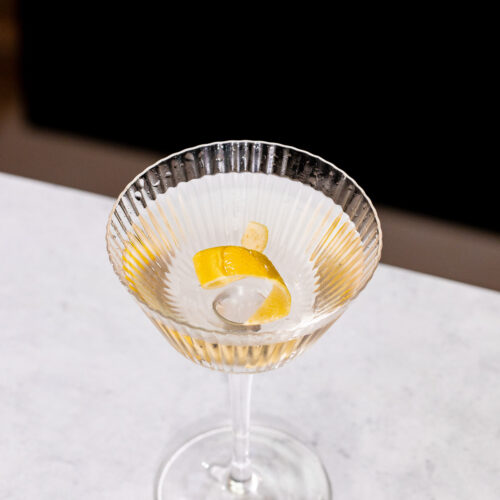 A clear Vesper martini in a ribbed martini glass with a lemon twist sits on a white surface.