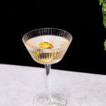 A Vesper martini in a ribbed glass coupe with a lemon twist sits on a light gray surface against a black background.