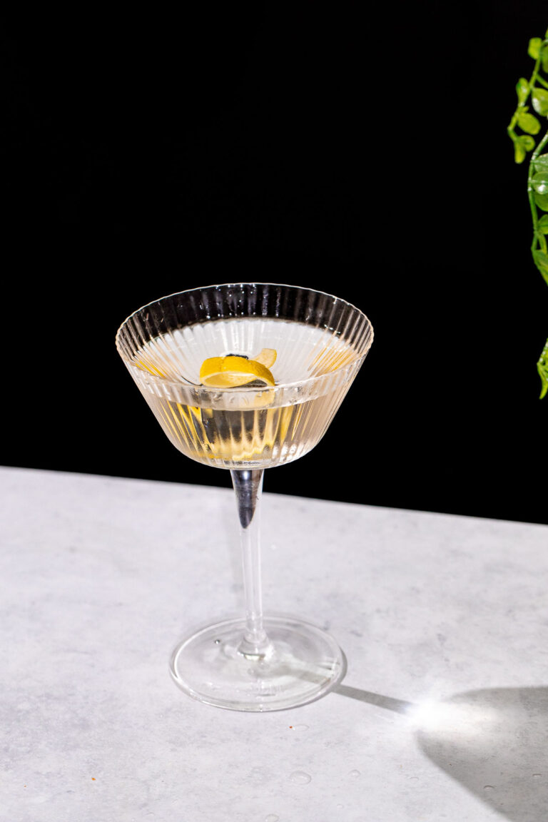 A Vesper martini in a ribbed glass coupe with a lemon twist sits on a light gray surface against a black background.