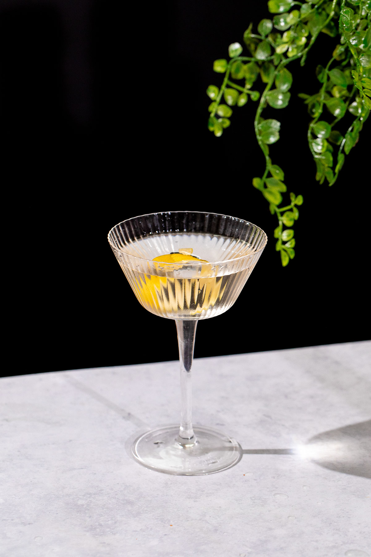 A Vesper martini with a lemon twist sits in a ribbed glass on a white surface, framed by green leaves in the background.