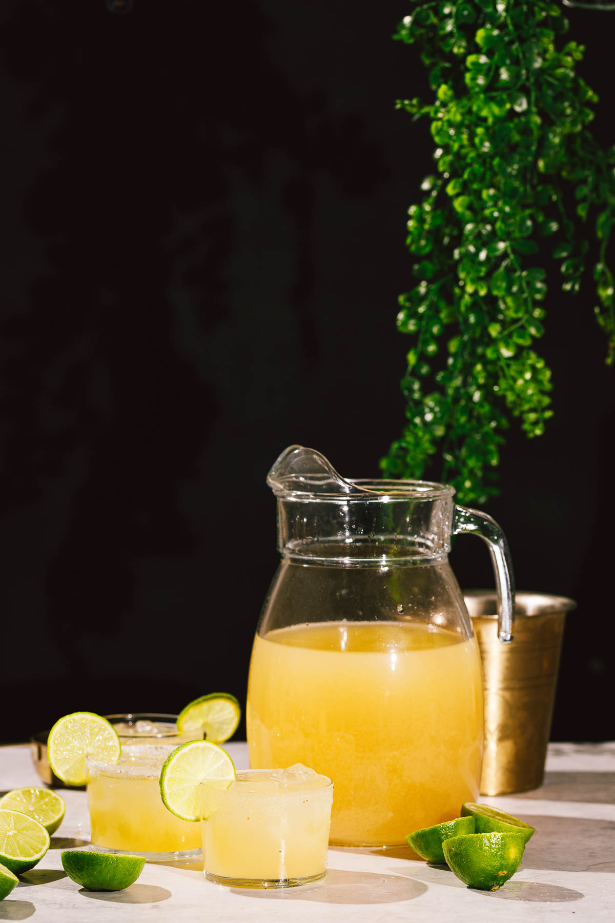 A glass pitcher and two lime-garnished glasses of margaritas sit on a table with limes and greenery.