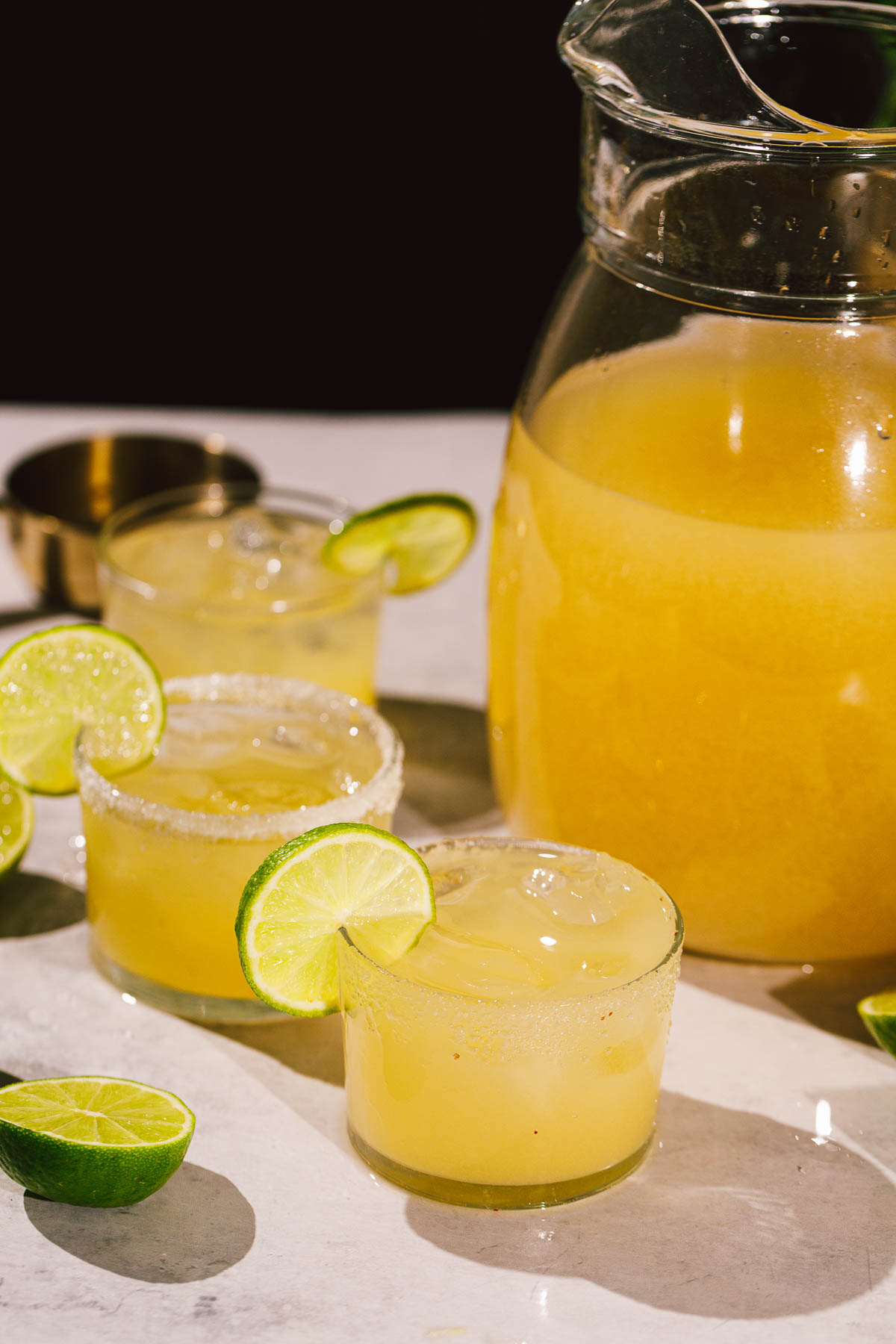 A pitcher and three ice-filled margarita with lime slices sit on a light surface with extra lime wedges.