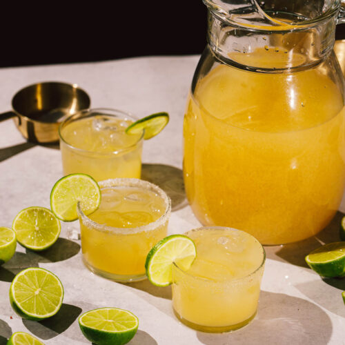 A pitcher and three glasses of margaritas with lime slices sit among halved limes on a white surface.