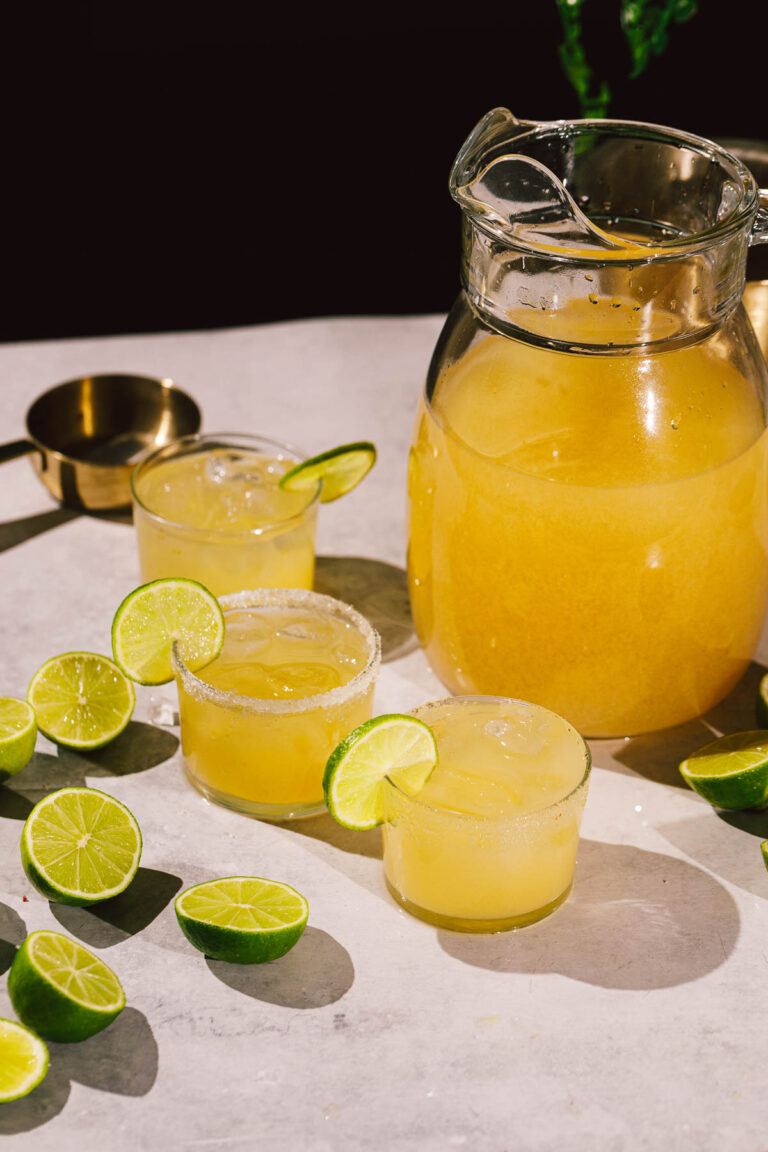 A pitcher and three glasses of margaritas with lime slices sit among halved limes on a white surface.