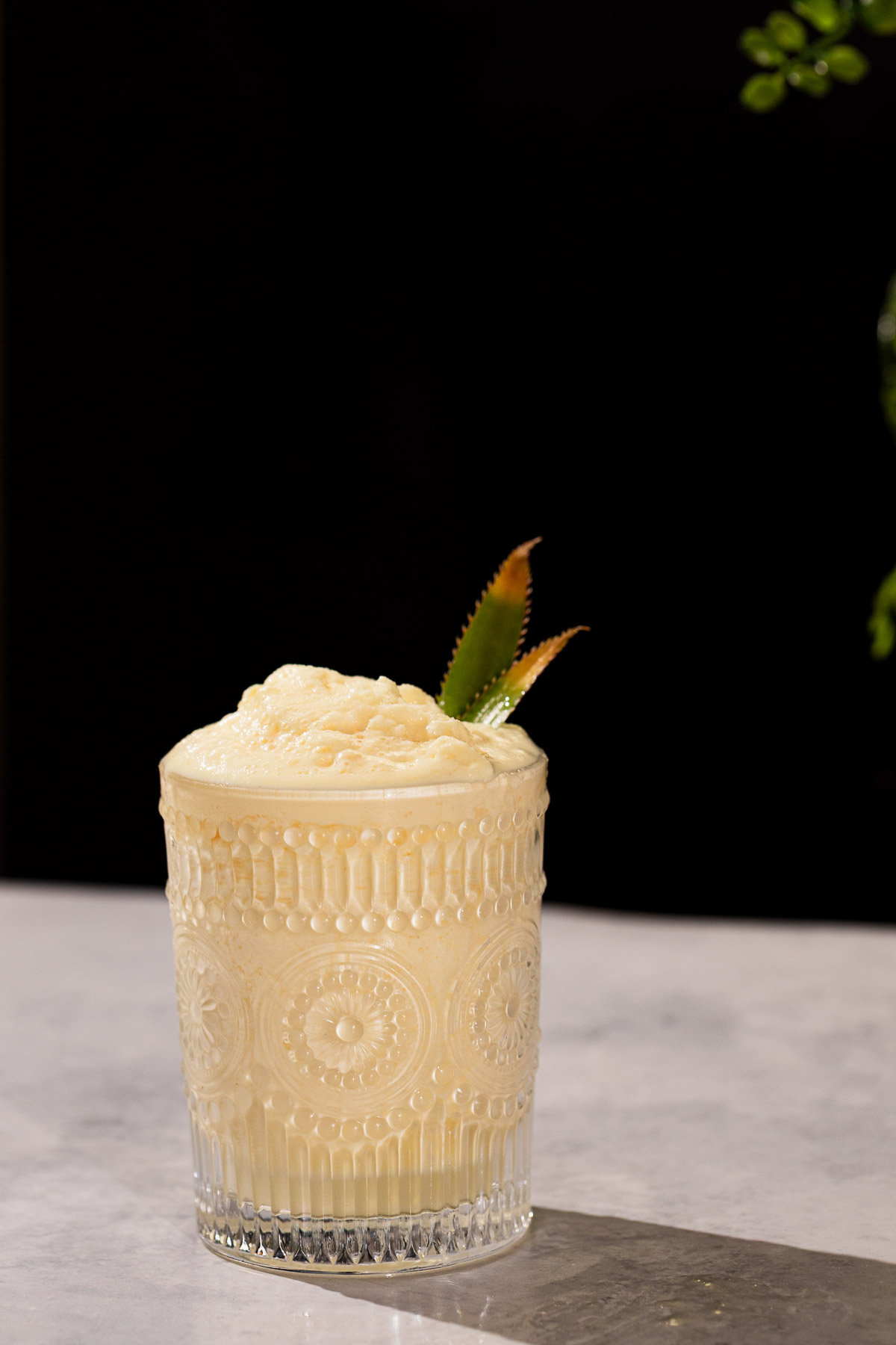 A glass of creamy batida frozen drink with two pineapple leaves on a marble surface against a dark background.