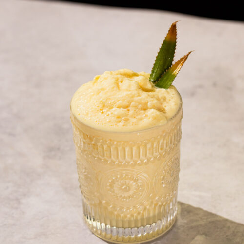 A glass of a frozen batida drink with two pineapple leaves sits on a light-colored surface.