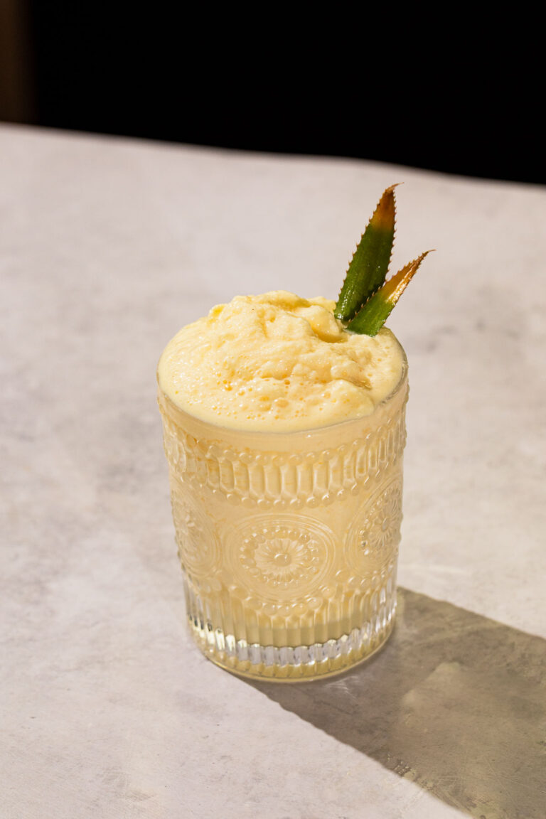A glass of a frozen batida drink with two pineapple leaves sits on a light-colored surface.