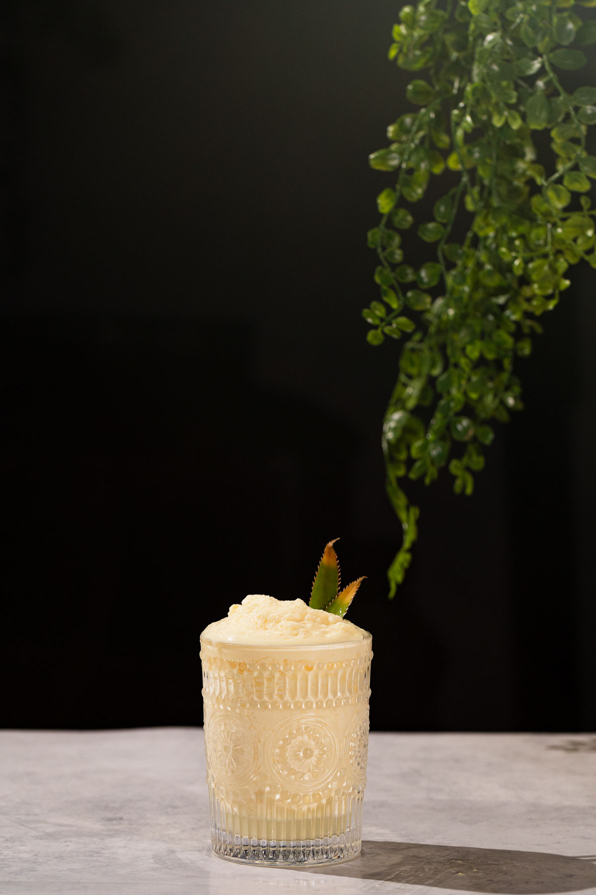 A batida drink with a pineapple leaf garnish sits on a gray surface beneath hanging green foliage.