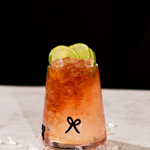 A glass with crushed ice, bramble cocktail, and three lime slices sits on a light surface with scattered ice pieces.