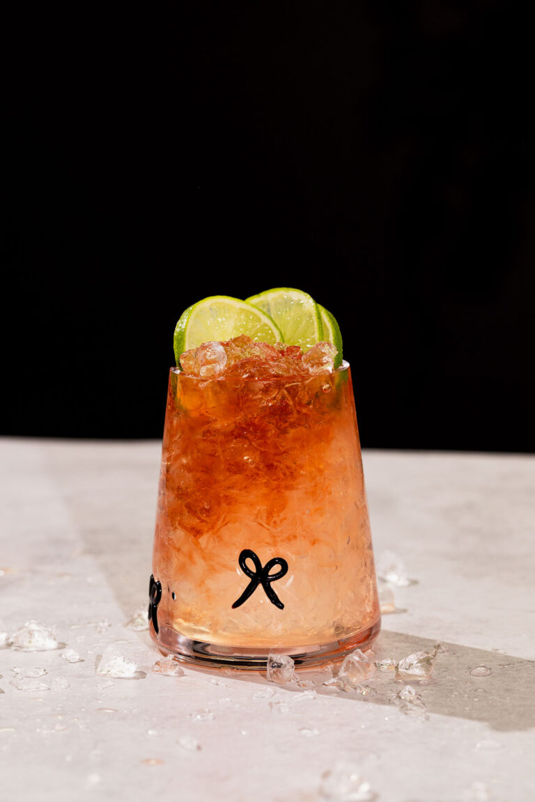 A glass with crushed ice, bramble cocktail, and three lime slices sits on a light surface with scattered ice pieces.