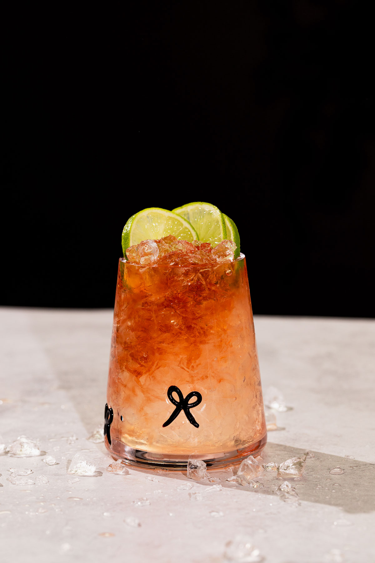 A glass with crushed ice, bramble cocktail, and three lime slices sits on a light surface with scattered ice pieces.