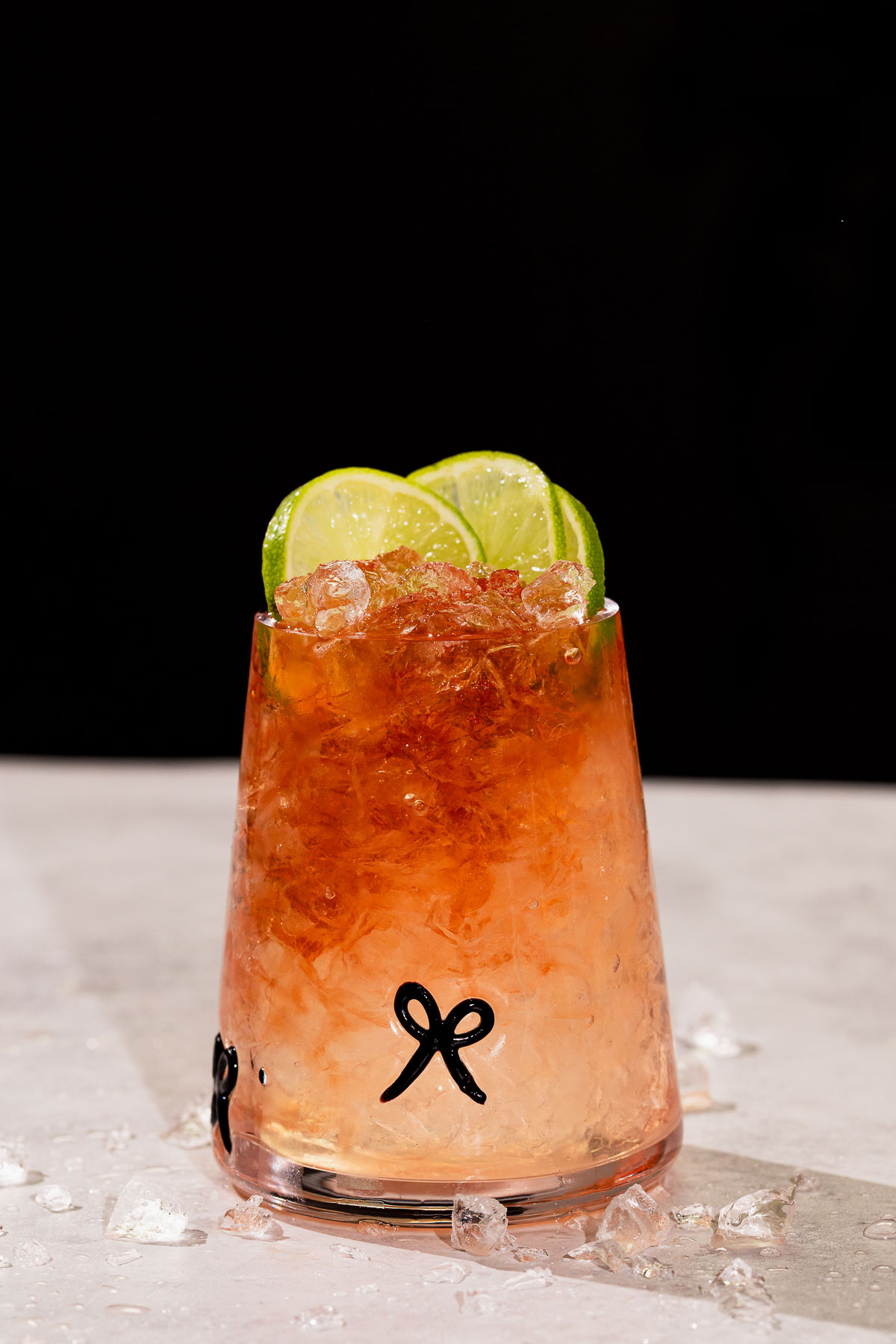 A bramble drink with crushed ice and three lime slices rests on a light surface scattered with ice.